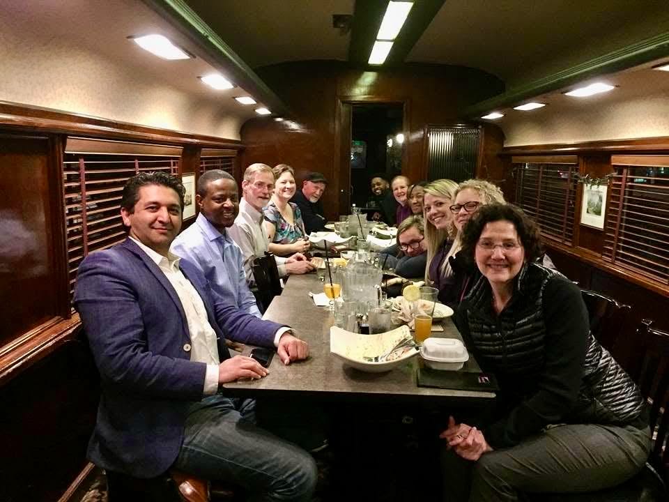 An Academy cohort seated along a long dinner table inside a rail car