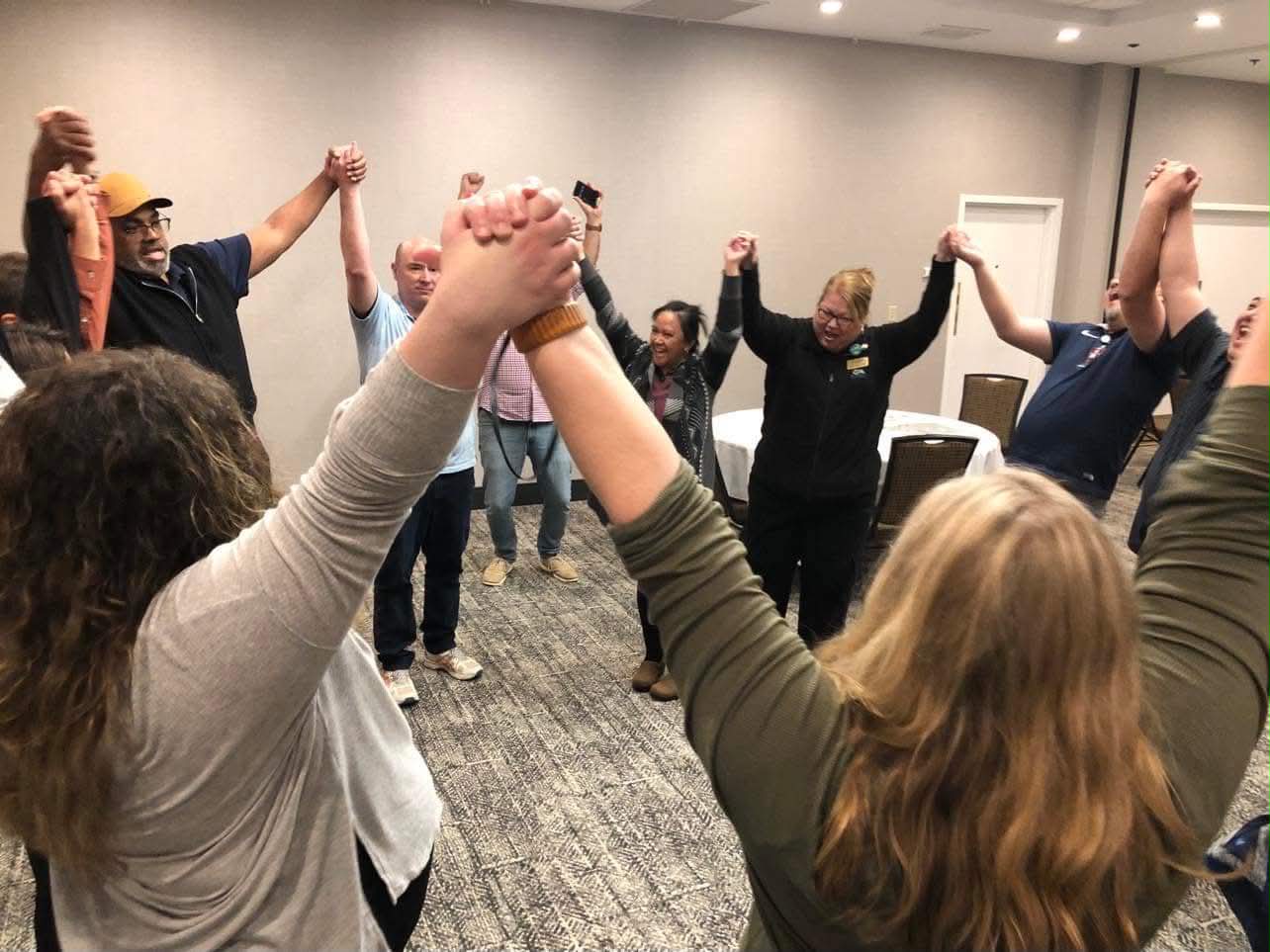 Workforce Deans Academy participants standing in a circle with hands joined and raised during an in-person session