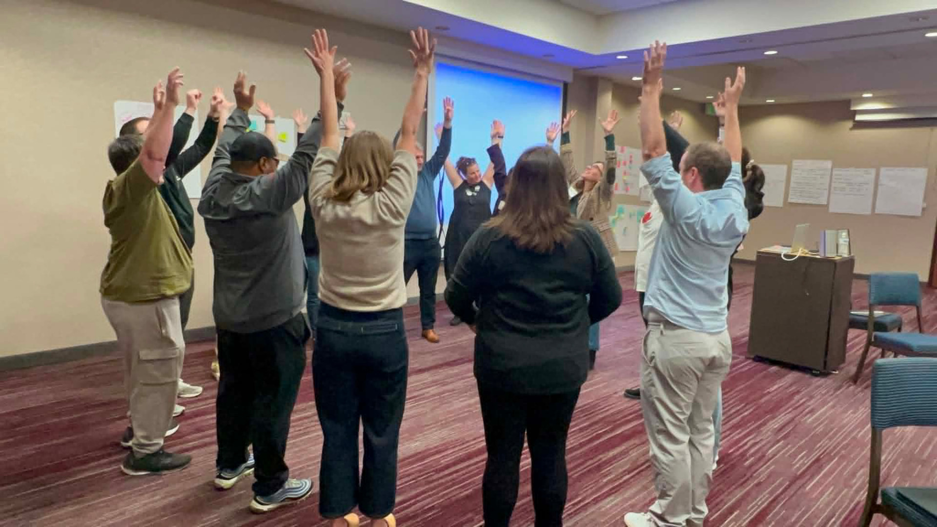 A full cohort standing with arms raised during a group energizer exercise